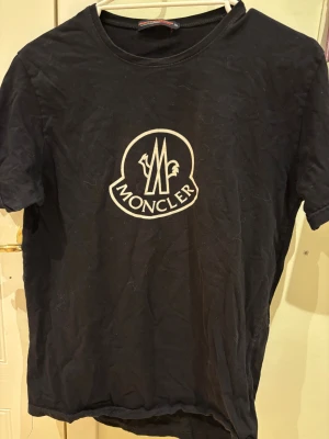 Svart Moncler t-shirt med logga - Svart t-shirt från Moncler med stor vit logga framtill och vertikal Moncler-text med logga på ryggen. Klassisk rund hals och korta ärmar. Tillverkad i mjuk bomull för en skön känsla och snygg passform.