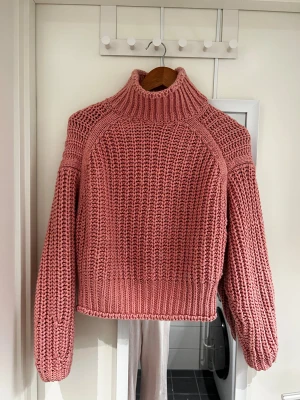 Rosa stickad polotröja chunky - Chunky stickad polotröja i en snygg rosa färg med bred ribbad krage och ballongärmar. Grovstickad struktur som ger en cool och mysig vibe. Perfekt för att hålla värmen och se trendig ut under höst och vinter.