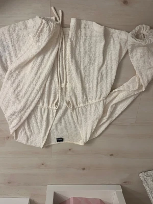 Vit omlott blus från Gina Tricot - Säljer en vit omlott blus från Gina Tricot med vågig struktur i tyget. Blusen har långa ärmar med resår vid ärmslut och knytband i midjan för en snygg passform. Perfekt för dig som gillar ljusa och luftiga toppar med coola detaljer.