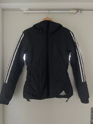 Svart Adidas pufferjacka med huva - Jättefin svart pufferjacka från Adidas med klassiska vita ränder längs ärmarna och logga på framsidan. Jackan har huva, dragkedja och justerbar nederkant med snörning. Storlek XS, knappt använd och i mycket fint skick