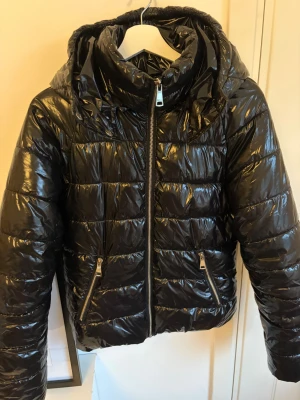 Svart glansig pufferjacka från Nelly - Säljer en svart, glansig pufferjacka från Nelly i storlek 36. Jackan har huva, två snedställda fickor med dragkedja och hel dragkedja framtill. Perfekt för kalla dagar och ger en cool vibe till din outfit.