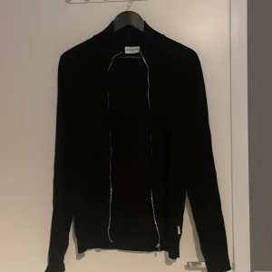 Svart full zip tröja från Lindbergh - Svart full zip tröja från Lindbergh i storlek S. Tröjan har en stilren design med hel dragkedja framtill och långa ärmar. Perfekt för dig som gillar minimalistisk stil och vill ha något som funkar till många olika outfits.