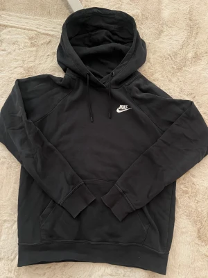 Svart Nike hoodie - Svart hoodie från Nike i storlek XS med klassisk logga broderad i vitt på bröstet. Tröjan har huva med snörning, känguruficka och långa ärmar. Mjuk och skön bomullsmix som passar perfekt när du vill vara chill. Snygg och enkel stil som funkar året runt.