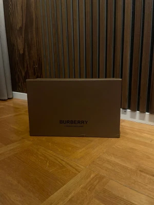 Burberry sneakers med rutig detalj - Snygga Burberry sneakers med svart skinn och mocka framtill, vit platt sula och klassisk beige-rutig textil på sidorna. Skorna har svarta snören och en stilren, modern look som sticker ut med Burberrys ikoniska mönster. Perfekt för dig som gillar exklusiva märken och streetstyle.
