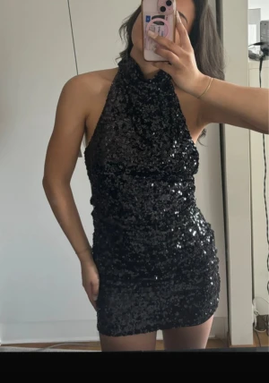 Svart glittrig paljettklänning - Säljer en svart kort paljettklänning med halterneck och öppen rygg. Klänningen är täckt av glittriga paljetter som ger en riktigt snygg och festlig look. Modellen är tight och ärmlös, perfekt för dig som vill sticka ut.