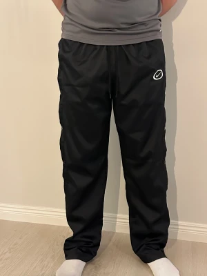 Svarta byxor/mjukis från Nike - Svarta träningsbyxor från Nike med knappar längs sidorna och elastisk midja med snörning. Byxorna har ett vitt Nike-logotyptryck på framsidan och är gjorda i lätt polyester som andas. Perfekta för sport eller chill. Snygg loose passform.