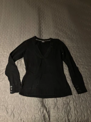 Svart stickad tröja från Esprit - Svart stickad tröja från Esprit med djup v-ringning och ribbade muddar. Långa ärmar med tre dekorativa knappar vid ärmslut och bred ribbad kant nedtill. Perfekt för dig som gillar enkel och stilren design.