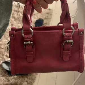 Rosa handväska med spänndetaljer - Snygg rosa handväska i skinn med coola metallspännen och ringar på handtagen. Väskan har en praktisk dragkedja upptill och en boxig form som ger den en trendig vibe. Perfekt storlek för det viktigaste och passar till många olika looks.