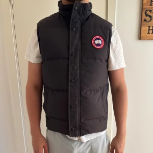 Svart dunväst från Canada Goose M - Svart dunväst från Canada Goose med hög krage och tryckknappar framtill. Klassisk rund logotyp-patch på bröstet. Västen har två sidofickor och är quiltad för extra värme. Perfekt för lager-på-lager under kyliga dagar.