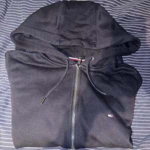 Svart hoodie från Tommy Hilfiger L - Svart hoodie från Tommy Hilfiger i storlek L. Klassisk design med dragkedja, huva med snörning och diskret logga på bröstet. Tillverkad i mjuk bomull och har ribbade muddar samt fickor framtill. Perfekt för en chill och stilren look.