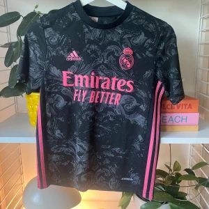 Real Madrid svart fotbollströja Adidas - Svart Real Madrid fotbollströja från Adidas med grått mönster och rosa detaljer. Tröjan har korta ärmar, rosa Adidas-logga, klubbmärke och sponsortryck framtill samt rosa ränder längs sidorna. Tillverkad i lätt och ventilerande polyester (AEROREADY).