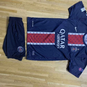 PSG matchställ med D. Doué #14 - Paris Saint-Germain matchställ med tröja och shorts. Tröjan är mörkblå med röd och vit rand i mitten, PSG-logga, Qatar Airways-tryck och Nike-märke. Nummer 14 och D. Doué på ryggen. Korta ärmar, snabbtorkande material, officiella Champions League och Respect-patchar.