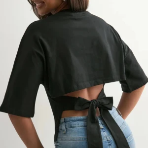 Tie Tee Svart Nelly  - Säljer en svart topp med halvlånga ärmar och snygg omlottdesign i ryggen som knyts med en stor rosett. Toppen är croppad och har en stilren look framtill, perfekt till jeans eller kjol. Ord.pris 250