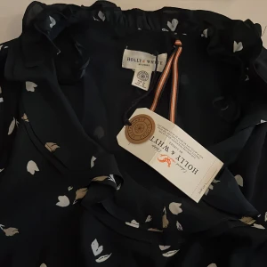 Mörkblå blommig klänning Holly & White - Supersöt mörkblå klänning från Holly & White med vita och beige blommor. Klänningen har volang vid halsen, lång ärm och knyt i midjan för en snygg siluett. Perfekt för dig som gillar en feminin och lekfull stil.