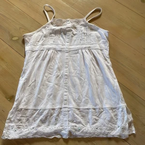 Vitt linne - Super sött vitt linne med tunna axelband, knappar framtill och broderade spetsdetaljer upptill och längs fållen. Storlek 146/152 dvs likt en xxs!☺️☺️