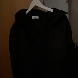 Svart softshelljacka från Stone Island - Svart softshelljacka från Stone Island med ikonisk kompasspatch på ärmen. Jackan har huva och är tillverkad med e.dye® teknologi för extra slitstyrka och vattenresistens. Perfekt för dig som gillar stilren och funktionell design.