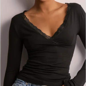Svart omlottopp med spetsdetalj - Svart omlottopp med lång ärm och v-ringad hals. Spetsdetaljer längs halsringningen ger en extra touch. Toppens passform är tight och materialet känns mjukt mot huden, perfekt för dig som gillar en stilren look med lite edge.