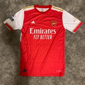 Arsenal Saka #7 matchtröja Adidas - Arsenals hemmatröja med Saka #7 på ryggen, tillverkad av Adidas i lätt och ventilerande material. Röd kropp med vita korta ärmar, guldiga detaljer och zigzag-mönster. Broderat klubbmärke och Adidas-logga framtill, samt sponsortryck. Perfekt för dig som älskar fotboll och Premier League.