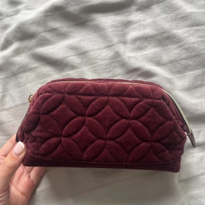 Vinröd quiltad necessär i sammet - Vinröd quiltad necessär i mjuk sammet med snyggt geometriskt mönster. Väskan har dragkedja upptill och är rymlig inuti, perfekt för smink eller småprylar. Har aldrig använt!