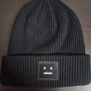 Svart ribbstickad mössa  - Svart ribbstickad mössa med en smiley framtill. 100% akryl. Använd ett par gånger men fortfarande i mycket gott skick.