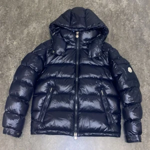 Moncler Maya mörkblå dunjacka - Säljer en Moncler Maya dunjacka i mörkblått med glansig finish. Jackan har huva, tvåvägsdragkedja, två snedställda fickor med dragkedja och Moncler-logga på ärmen. Insidan är brun och jackan är riktigt puffig och varm – perfekt för kalla dagar.