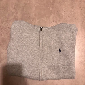 Grå hoodie från Polo Ralph Lauren - Snygg grå hoodie från Polo Ralph Lauren med dragkedja och klassisk blå logga på bröstet. Tröjan har huva, känguruficka och är fodrad med mjuk fleece på insidan. Perfekt för chill dagar och enkel att matcha med jeans eller joggers. Tröjan har inte blivit använt så ofta utan har vart i garderoben då den inte passar mig riktigt 