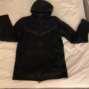 Svart Nike hoodie - Svart hoodie från Nike i storlek S med hel dragkedja och huva. Tröjan har diskreta svarta detaljer över bröstet och en ficka med dragkedja på ärmen. 