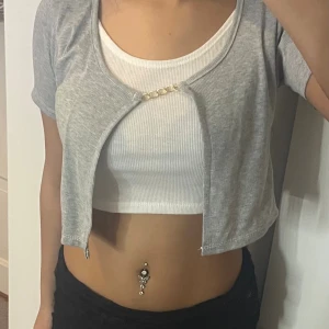 Grå croppad bolerotopp med kedjedetalj - Säljer en grå croppad bolerotopp med korta ärmar och en snygg guldfärgad kedjedetalj framtill. Under finns en vit ribbad linnetopp som ger en fräsch lager-på-lager-look. Perfekt för att lyfta en enkel outfit med lite extra edge.