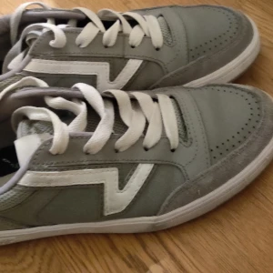 Gråa Vans sneakers med vit detalj - Säljer ett par gråa Vans sneakers med vit sula och vit sidodetalj. Skorna har snörning, rund tå och är tillverkade i en mix av mocka och syntetmaterial. Klassisk Vans-yttersula och stilren design som passar till det mesta.