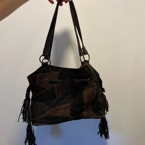 Patchwork axelväska i mocka med fransar - Säljer en brun patchwork axelväska i mjuk mocka med coola fransdetaljer på sidorna. Väskan har snörning upptill, dragkedjeficka på baksidan och rejäla axelband i samma material. Perfekt för dig som gillar en bohemisk och unik stil.