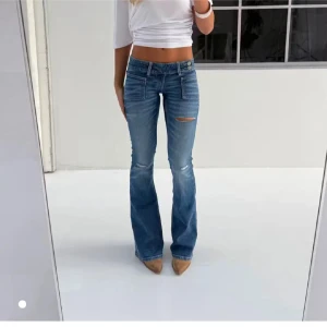 Blå bootcut jeans med slitning - Snygga blå bootcut jeans med lätt slitning och en liten reva på låret. Jeansen har klassiska fickor fram och bak, låg midja och är tillverkade i ett mjukt denimtyg. Perfekta för dig som gillar en avslappnad men trendig look.