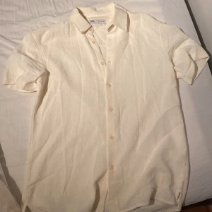 Vit/sand kortärmad skjorta från Zara - Snygg vit/sand färgad skjorta från Zara i storlek M. Skjortan har korta ärmar, klassisk krage och knappar framtill. Materialet är mjukt och strukturerat, vilket ger en chill och modern vibe. Perfekt för varmare dagar och enkel att styla med jeans eller shorts.