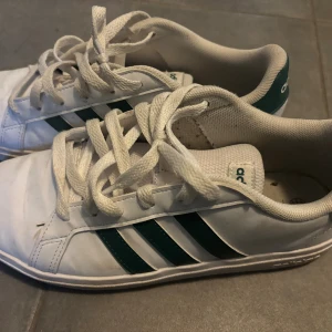 Vita Adidas sneakers med gröna ränder - Klassiska vita Adidas sneakers med gröna ränder på sidorna och vit sula. Skorna har snörning och är tillverkade i syntetmaterial med textilfoder. 