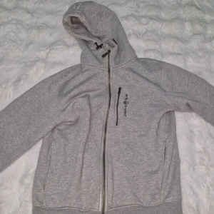 Grå hoddie från Sail Racing med dragkedja - Snygg ljusgrå hoodie från Sail Racing med hel dragkedja framtill, broderad logga på bröstet och huva med snörning. Tröjan har en extra dragkedjeficka på bröstet och är tillverkad i mjuk bomullsblandning. Perfekt för chill dagar eller när du vill vara casual.