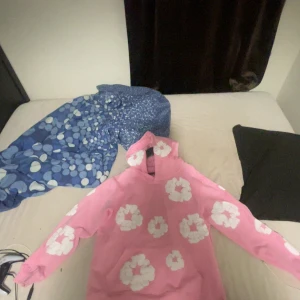 Rosa hoodie med vita blommor Denim Tears - Säljer en rosa hoodie från Denim Tears med stora vita blommor tryckta över hela tröjan. Hoodien har en klassisk känguruficka och huva, perfekt för dig som vill sticka ut med en färgglad och unik stil. Materialet är mjukt och skönt, passar perfekt till chill dagar.