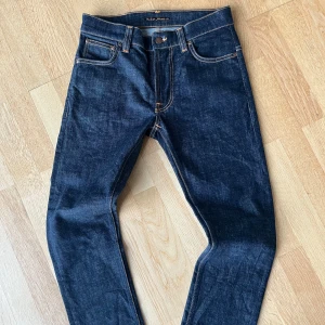 Mörkblå jeans från Nudie Jeans - Snygga mörkblå jeans från Nudie Jeans med klassisk orange söm och ikonisk broderad bakficka. Jeansen har rak passform, fem fickor och är tillverkade i robust denim av bomull. Perfekta för dig som gillar stilrena och tidlösa jeans.