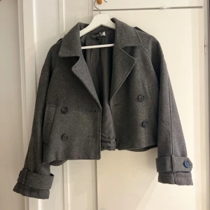 Grå croppad  från H&M - Snygg grå croppad jacka från H&M med bred krage och dubbelknäppning. Jackan har långa ärmar med dekorativa knappar vid ärmslut. Perfekt för dig som gillar en stilren och trendig look.