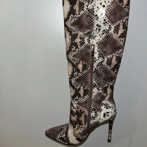 Höga boots med ormskinnsmönster - Säljer ett par höga boots med spetsig tå och stilettklack i ormskinnsmönster. Färgerna går i beige och brunt med dragkedja på sidan. Förövrigt helt oanvända i storlek 7 (40) jag har dock 39 och de passar mig så mer av en 39 1/2