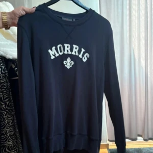 Mörkblå sweatshirt från Morris - Säljer en mörkblå sweatshirt från Morris med vit logga och emblem framtill. Tröjan har rund halsringning och långa ärmar, perfekt för en chill och stilren look.