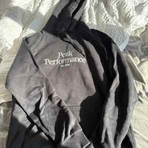 Svart hoodie från Peak Performance - Svart hoodie från Peak Performance med vit broderad logga på bröstet. Klassisk modell med huva och snörning samt känguruficka framtill. Mjuk insida och ribbade muddar vid ärmslut och nederkant. Perfekt för chill dagar eller när du vill vara extra bekväm.
