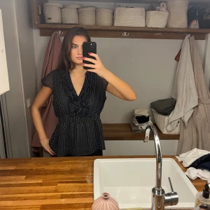 Svart prickig blus med volang H&M - Supersöt svart blus från H&M med vita prickar, v-ringning och volangdetaljer längs ringningen. Blusen har korta ärmar i transparent tyg och smockad midja som ger en snygg siluett. Perfekt för dig som gillar feminina detaljer och mönster. Storlek XL men jag är själv S och har använt denna. 