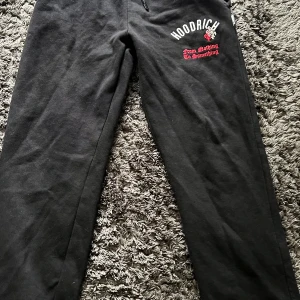 Svarta Hoodrich mjukisbyxor - Svarta mjukisbyxor från Hoodrich med broderad logga och texten 'From Nothing To Something' i rött och vitt på ena benet. Sköna och avslappnade med normal passform, perfekta för chill eller streetwear. Materialet är mjukt och bekvämt bomullstyg.