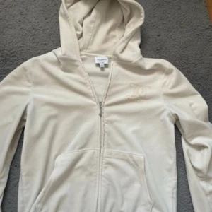 Beige Juicy Couture hoodie med dragkedja - Säljer en beige hoodie från Juicy Couture med klassisk logga på bröstet och stor Juicy Couture-text på ryggen. Tröjan har huva, fickor fram och dragkedja. Mjuk velourkänsla och långärmad modell, perfekt för chill dagar. Den är använd ganska så mycket så färgen har gått bort lite där fram men annars super fin på!💞