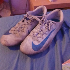 Nike fotbollsskor i vitt och blått - Säljer ett par Nike fotbollsskor i vitt med blå detaljer och snörning. Skorna har en låg profil och är tillverkade i syntetmaterial med klassisk Nike swoosh på sidan. Perfekta för dig som vill ha stilrena och lätta skor på planen.
