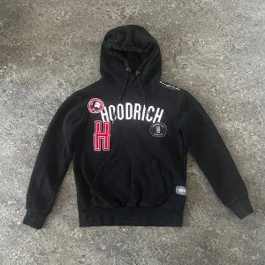 Hoodrich hoodie - Svart hoodie från Hoodrich med broderade patchar och tryck framtill. Tröjan har huva med snörning, magficka och ribbade muddar. Stora vita och röda detaljer på bröstet.