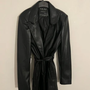 Svart skinn kappa från Amisu - Säljer en svart  skin kappa / jacka i skinnimitation från Amisu. Jackan har breda slag, knytskärp i midjan och två fickor framtill.  Storlek 34. Mycket bra skick . 