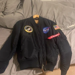 NASA bomberjacka från Alpha Industries - Säljer en mörkblå bomberjacka från Alpha Industries med NASA-patch, amerikansk flagga och coola detaljer som 'Remove Before Flight'-tag. Jackan har orange foder, dragkedja och flera broderade märken. Perfekt för dig som gillar rymd vibes och streetstyle.