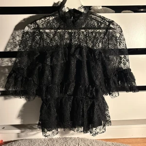 Svart spetsblus med volanger Bubbleroom - Säljer en svart blus från Bubbleroom i storlek 36. Blusen är gjord i transparent spets med flera lager volanger och har en hög hals samt korta ärmar med volangkant. Andvänt 2 gånger 