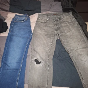 Blå och grå jeansbyxor - Två par jeans, ett par klassiskt blå jack and jones och ett par grå nudie med skön slitning på ena benet. Båda har femficksdesign och raka ben. De blå är i en djup blå nyans medan de grå har en tvättad look. Perfekta för en avslappnad stil. Grå 31/30, blå 29/30 har av er vid frågor!