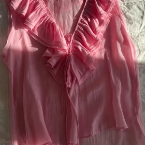Rosa volangblus från Zara - Supersöt rosa blus från Zara med volangdetaljer längs v-ringningen och knyt framtill. Tunn och luftig modell som är perfekt för varmare dagar. Ärmlös och i ett lätt, skönt material som ger en flowy känsla.
